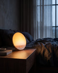 Aetheris Sunrise Lamp glowing warm on a wooden nightstand at dawn, LED clock showing 06:00